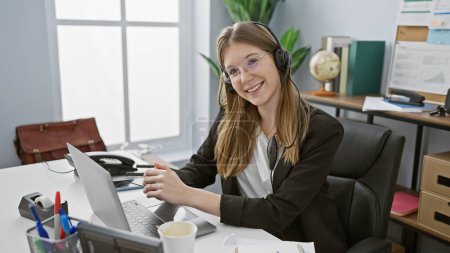 Photo for Smiling young adult caucasian woman in headset at office with laptop and coffee - Royalty Free Image