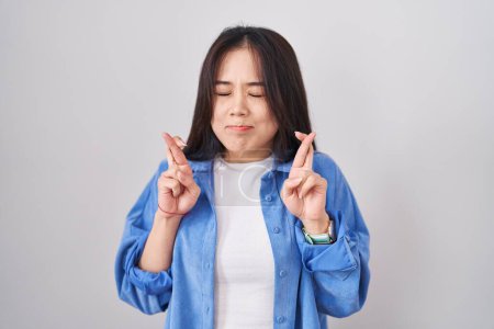 Téléchargez les photos : Jeune femme chinoise debout sur fond blanc geste doigt croisé souriant avec espoir et les yeux fermés. chance et concept superstitieux. - en image libre de droit