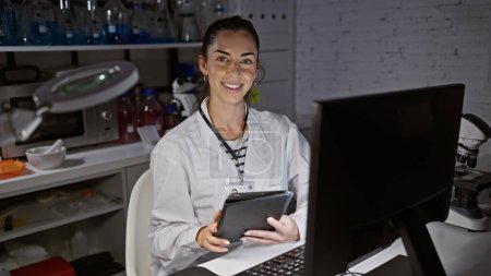 Photo for Smiling attractive young hispanic woman scientist, radiating with beauty, engages with touchpad and computer at her laboratory workspace, immersed in her night-time research. - Royalty Free Image