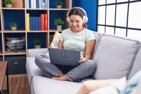 Téléchargez les photos : Jeune femme hispanique utilisant un ordinateur portable à la maison portant des écouteurs regardant positif et heureux debout et souriant avec un sourire confiant montrant les dents - en image libre de droit