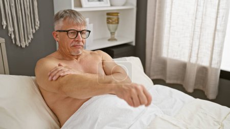Photo for Mature shirtless man with glasses seated in a white, well-lit bedroom looking thoughtful - Royalty Free Image