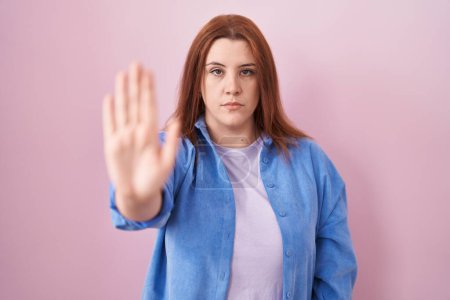 Mujer hispana joven con el pelo rojo de pie sobre fondo rosa haciendo dejar de cantar con la palma de la mano. expresión de advertencia con gesto negativo y grave en la cara. 