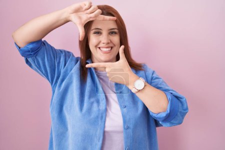 Téléchargez les photos : Jeune femme hispanique aux cheveux roux debout sur fond rose souriant faisant cadre avec les mains et les doigts avec le visage heureux. concept de créativité et photographie. - en image libre de droit