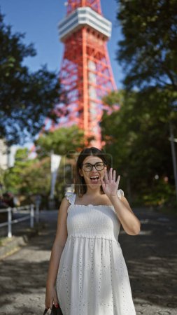 Smiling, glasses-clad beautiful hispanic woman cheerfully waving hello by tokyo tower, a famous japanese landmark with a grand cityscape view.