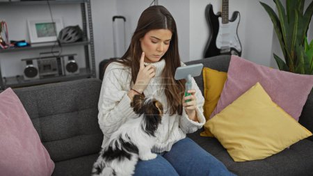 Eine junge hispanische Frau erwägt die Verwendung eines Haustier-Haarentferners, während sie mit ihrem schönen Hund in einem gemütlichen Wohnzimmer sitzt.