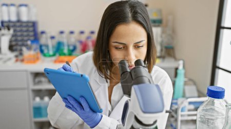 Téléchargez les photos : Une jeune scientifique hispanique analysant des échantillons au microscope tout en tenant un comprimé dans un cadre de laboratoire. - en image libre de droit