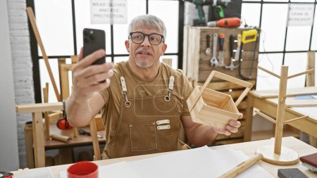Téléchargez les photos : Homme aux cheveux gris dans un atelier tenant un objet en bois et prenant selfie avec smartphone - en image libre de droit