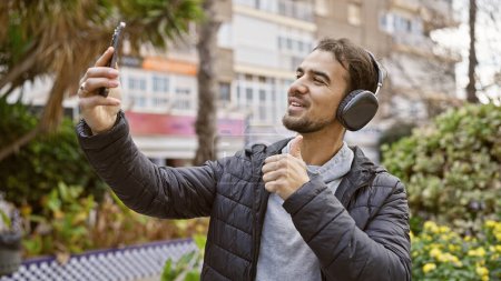 A smiling young man takes a selfie with a smartphone in an urban park, giving a thumbs up while wearing headphones.