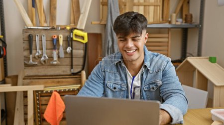 Photo for Handsome young hispanic man, a professional carpenter, cheerfully working with laptop at carpentry workshop - Royalty Free Image