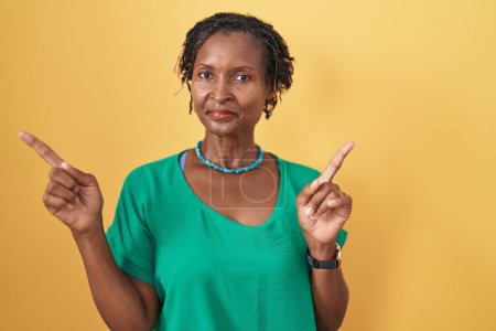 Foto de Mujer africana con rastas de pie sobre fondo amarillo sonriendo confiada señalando con los dedos a diferentes direcciones. espacio de copia para publicidad - Imagen libre de derechos