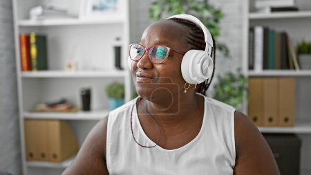 Foto de Mujer de negocios afroamericana sonriente, una exitosa gerente que piensa ideas en el trabajo, escuchando su canción favorita, mirando a un lado en la oficina. - Imagen libre de derechos