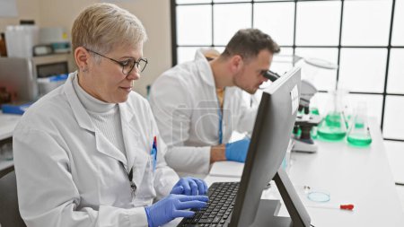 Foto de Una mujer y un hombre con batas de laboratorio trabajan diligentemente en un laboratorio brillante, centrándose en la investigación informática y el análisis científico. - Imagen libre de derechos