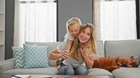 Relaxed caucasian mother sitting comfortably on sofa at home, holding her little daughter playfully on her back, the family radiating happiness
