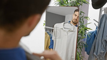 Photo for A handsome young hispanic man in a clothing room, contemplating a shirt while looking in the mirror reflecting his beard and stylish demeanor. - Royalty Free Image