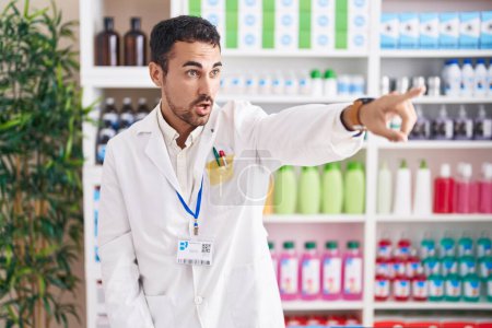 Téléchargez les photos : Bel homme hispanique travaillant à la pharmacie pharmacie pointant du doigt surpris devant, bouche ouverte expression étonnée, quelque chose sur le devant - en image libre de droit