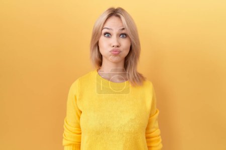Foto de Mujer caucásica joven con suéter amarillo soplando mejillas con cara divertida. boca hinchada de aire, expresión loca. - Imagen libre de derechos