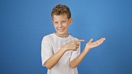 Photo for Adorable blond boy all smiles, confidently presenting and pointing to copy space on isolated blue background. joyful expression of this little caucasian kid standing, enjoying the show. - Royalty Free Image