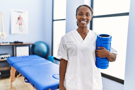 Téléchargez les photos : Femme afro-américaine physiothérapeute souriant confiant tenant rouleau de mousse à la clinique de réadaptation - en image libre de droit