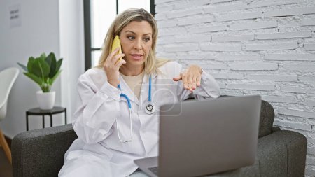 Foto de Joven doctora rubia usando laptop hablando en smartphone en la clínica - Imagen libre de derechos