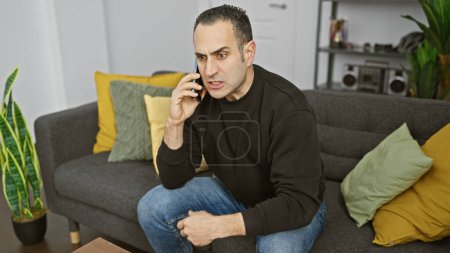 Téléchargez les photos : Un homme hispanique regardant préoccupé tout en parlant sur un smartphone dans un cadre de salon moderne - en image libre de droit