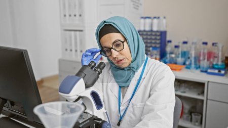 Téléchargez les photos : Une femme scientifique mature dans un hijab examine des échantillons au microscope dans un cadre de laboratoire, transmettant le professionnalisme et la concentration. - en image libre de droit