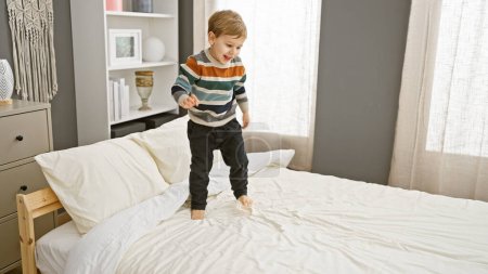 Photo for Happy toddler boy in striped sweater jumps on a clean bed in a cozy bedroom interior. - Royalty Free Image