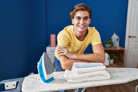 Foto de Joven hombre caucásico sonriendo confiado apoyado en tabla de planchar en la sala de lavandería - Imagen libre de derechos