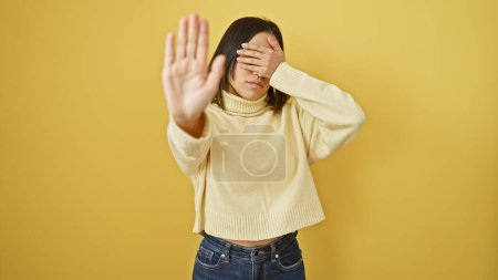 A young hispanic woman in a cream sweater covers her face against a yellow background, symbolizing avoidance or privacy.