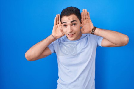 Photo for Young hispanic man standing over blue background trying to hear both hands on ear gesture, curious for gossip. hearing problem, deaf - Royalty Free Image