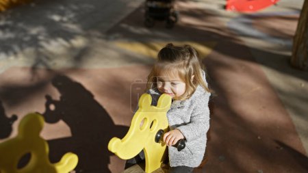 Cute blonde girl playing on a yellow animal-shaped playground spring rider in a sunny park.