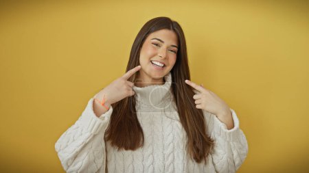 Photo for Happy young woman in a white sweater pointing at her smile against a vibrant yellow background, expressing joy. - Royalty Free Image