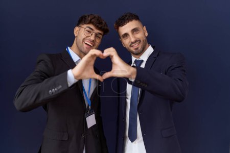 Téléchargez les photos : Deux hommes d'affaires hispaniques portant des vêtements d'affaires souriant en amour faisant forme de symbole de coeur avec les mains. concept romantique. - en image libre de droit