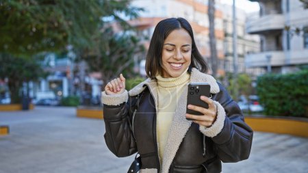Photo for A smiling young hispanic woman engaging with her smartphone on a sunny city street. - Royalty Free Image