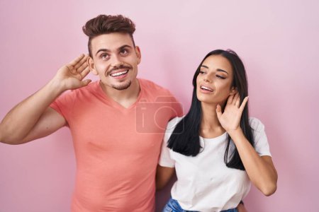 Foto de Joven pareja hispana de pie sobre fondo rosa sonriendo con la mano sobre el oído escuchando un rumor o chismes. concepto de sordera. - Imagen libre de derechos