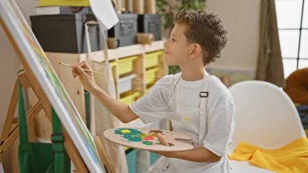 Téléchargez les photos : Adorable garçon artiste blond, souriant en toute confiance alors qu'il aime dessiner au studio d'art, immergé dans sa leçon de peinture - en image libre de droit