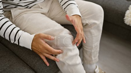 Photo for Hispanic man's hands gripping knee in pain, sitting on comfy living room sofa, suffering from leg joint injury at home, basking in sunlight yet unable to relax due to discomfort - Royalty Free Image