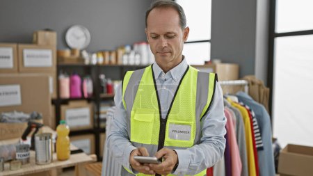 Photo for Concentrated middle age man donned in reflective vest, altruistically volunteering at local charity center, diligently typing message on his smartphone amidst bundles of donation boxes. - Royalty Free Image