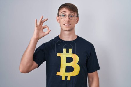 Foto de Hombre rubio caucásico con camiseta bitcoin sonriendo positiva haciendo signo de ok con la mano y los dedos. expresión exitosa. - Imagen libre de derechos