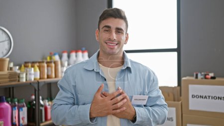 Heartwarming portrait of young, handsome hispanic man volunteering at charity center, chest out, hands on heart, standing tall, smiling in emotional service to his community