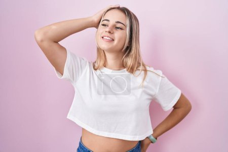 Téléchargez les photos : Jeune femme blonde debout sur fond rose souriant cheveux touchants confiants avec geste de la main vers le haut, posant attrayant et à la mode - en image libre de droit