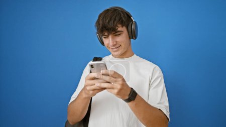 Photo for Cool young hispanic boy, a confident student, smiling while typing a text on his smart phone. standing casually, backpack on, listening to music with headphones on a isolated blue background. - Royalty Free Image