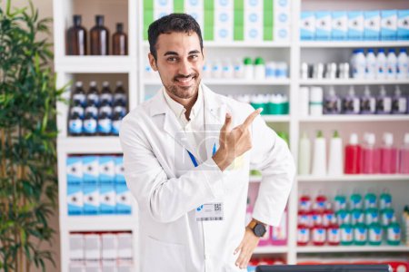 Téléchargez les photos : Bel homme hispanique travaillant à la pharmacie pharmacie gai avec un sourire sur le visage pointant avec la main et le doigt sur le côté avec une expression heureuse et naturelle - en image libre de droit