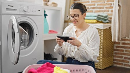 Foto de Joven hermosa mujer hispana haciendo la colada usando smartphone en la sala de lavandería - Imagen libre de derechos