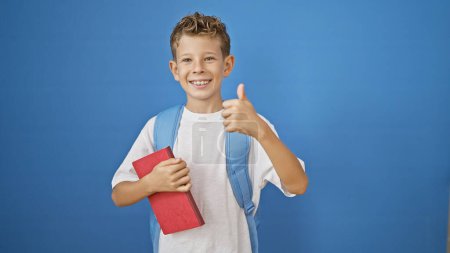 Photo for Adorable blond boy student, confidently holding book while giving thumb up ok gesture, smiling widely on isolated blue background, celebrating the joy of learning. - Royalty Free Image