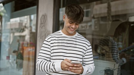 Téléchargez les photos : Joyeux jeune homme hispanique embrassant le bonheur en plein air, textant et souriant dans la rue urbaine à l'aide de son téléphone portable, illuminant la ville avec sa connexion au monde numérique - en image libre de droit
