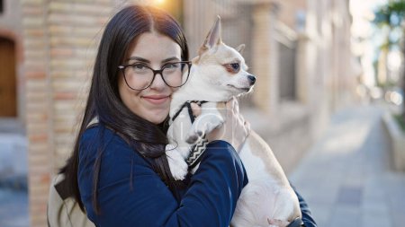 Foto de Mujer hispana joven con perro chihuahua vistiendo mochila sonriendo juntos en la calle - Imagen libre de derechos