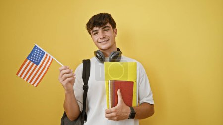 Téléchargez les photos : Jeune étudiant hispanique dynamique, un adolescent prometteur avec des livres et drapeau des États-Unis, debout avec confiance avec style de vie décontracté étalé, isolé sur un fond jaune vif. - en image libre de droit