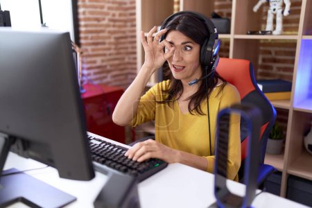 Photo for Middle age hispanic woman playing video games using headphones smiling happy doing ok sign with hand on eye looking through fingers - Royalty Free Image