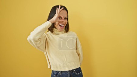 Téléchargez les photos : Une jeune femme joyeuse fait un geste "ok" près de son œil contre un mur jaune, exsudant charme et positivité. - en image libre de droit