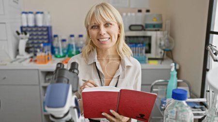 Foto de Mujer rubia científica en bata de laboratorio leyendo un cuaderno dentro de un entorno de laboratorio moderno. - Imagen libre de derechos